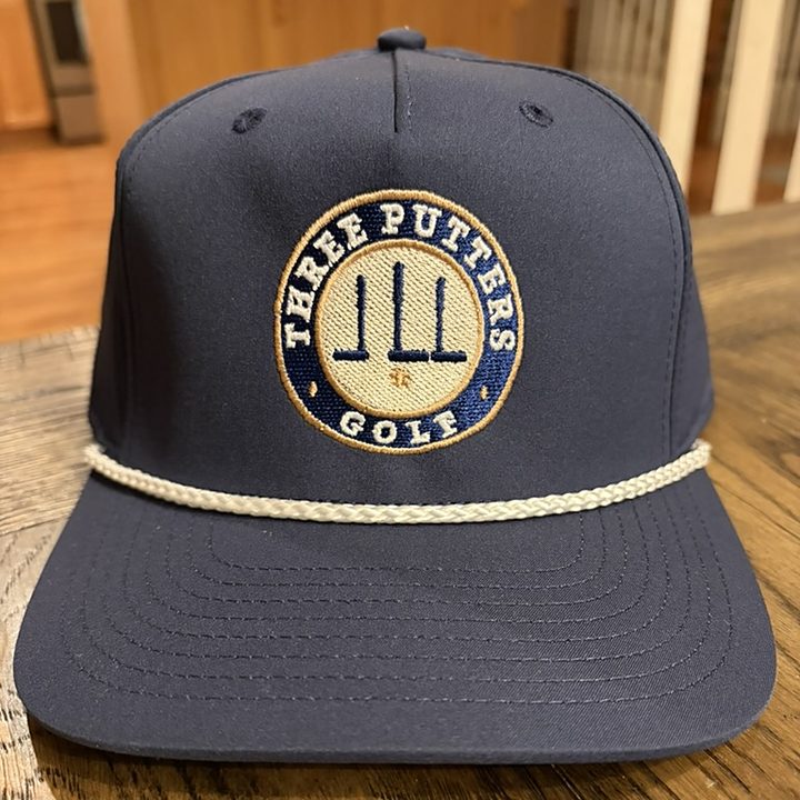 Club Seal Rope Hat — navy rope cap with the embroidered Three Putters Golf seal in navy, cream and gold, and a cream braided rope